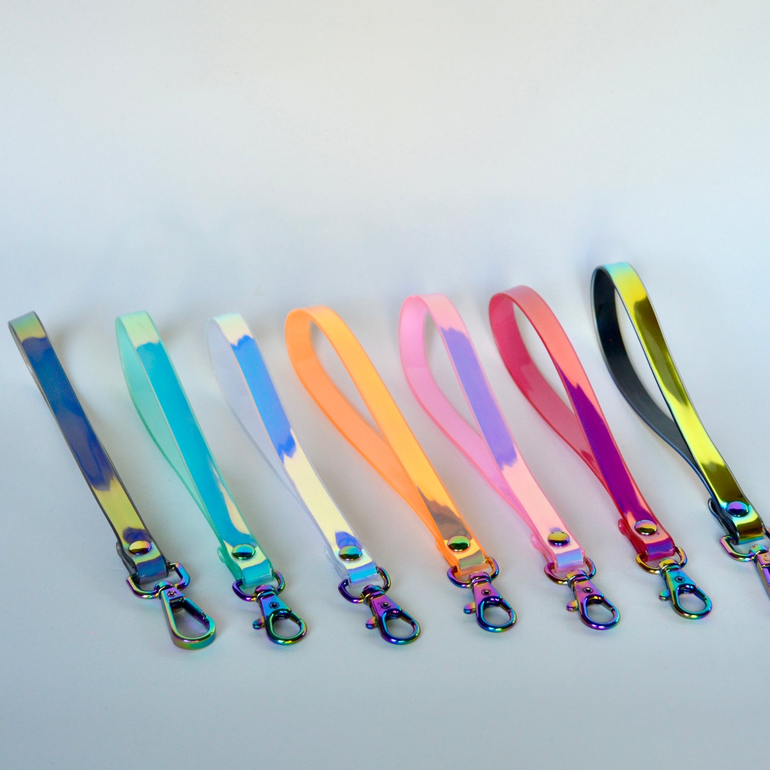 Six iridescent holographic wristlets in various colors, arranged in a row on a light gray background. Each wristlet features a flat strap with a metal lobster clasp at the end.